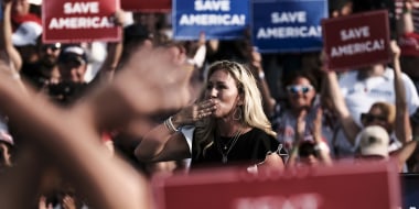Image: Marjorie Taylor Greene on a stage at a rally with people in the crowd holding signs that read,\"Save America\".
