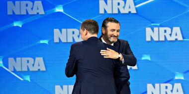 Image: Ted Cruz hugging someone on stage which has a background that has \"NRA\" repeated all over.