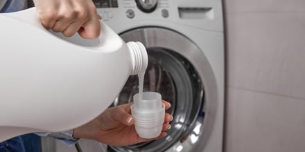 In bathroom where washing machine is located, girl pours the laundry conditioner into lid to give clean laundry pleasant smell and softness. From white plastic bottle, rinse is poured into the cap