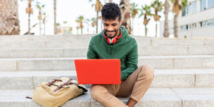 Portrait of hispanic young student working on laptop while sitting on steps outdoors - College, millennial people and education online concept