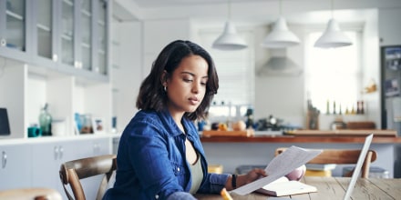 Shot of a young woman using a laptop and  going through paperwork while working from home