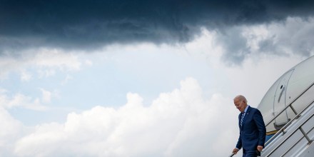 Image: President Joe Biden climbing down the stairs from a plane against a dark cloudy sky.