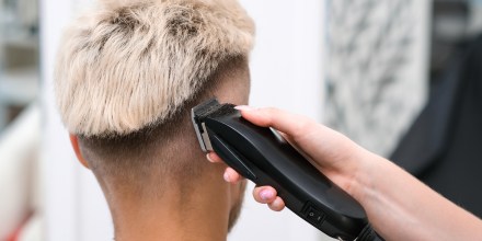 Haircut process of blond young man with hair trimmer in armchair in barbershop salon.
