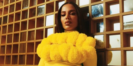 Anitta, in a yellow Aknvas sweater adorned with fuzzy chest patterns, sits in front of a mirrored cube-patterned wall.