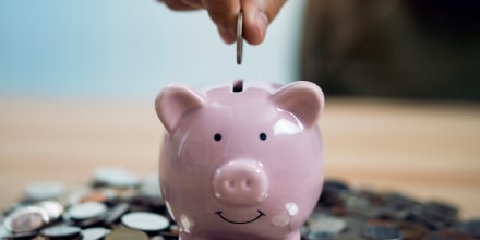 Person Putting Coin In Piggy Bank At Table