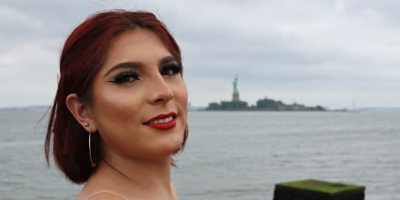 Michelle Carranza smiles outside, the statue of Liberty can be seen across the water in the distance