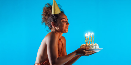 Profile view of happy young woman standing against blue background and blowing out candles on a birthday cake.