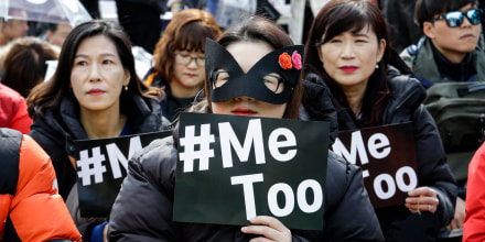 A person wears a black mask and holds a sign that says "#Me Too" in a crowd of protesters