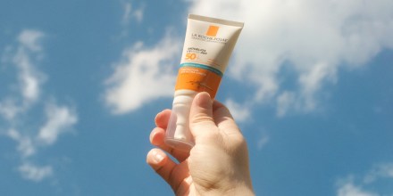 A hand holds tone bottle of sunscreen against a blue sky with clouds