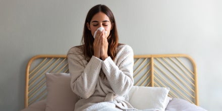 Young caucasian woman blowing nose with paper tissue wearing a robe sitting on bed suffering from a cold.