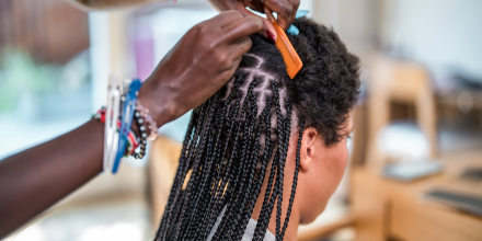 A mid adult Black woman braids the hair of a mixed race female in a domestic setting, showcasing a traditional hairstyle being made