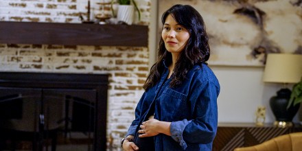 A woman stands in a living room and holds her pregnant belly