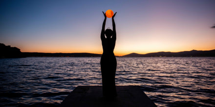 Silhouette of young woman in black dress, backdrop of twilight by sea, holding full moon in hands.