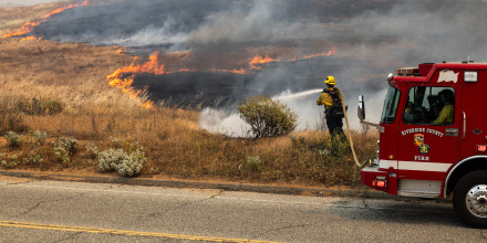 A firefighter standing in front of a fire truck aims a running hose at a fire overtaking a field by a road