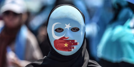 Image: A demonstrator wearing a mask painted with the colours of the flag of East Turkestan and a hand bearing the colours of the Chinese flag