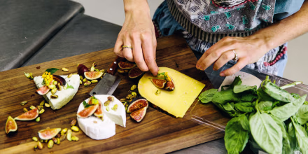 Woman preparing variety cheese plate with figs, nuts, pistachios, basil on wooden chopping board