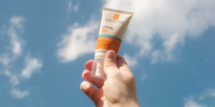 A hand holds tone bottle of sunscreen against a blue sky with clouds