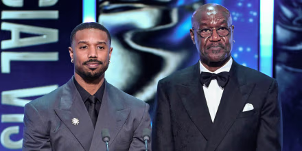 Michael B. Jordan and Delroy Lindo stand at a lectern on stage during an award ceremony. They look nonplussed.