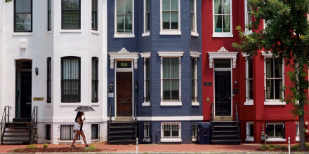 A woman with an umbrella walks past red, white and blue painted row houses.