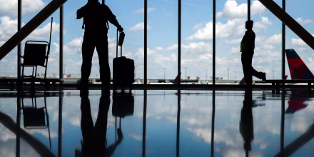 Silhouettes of people standing in an airport near large windows with a view of the sky and tarmac, reflections can be seen on the floor