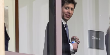 Sam Altman smiles toward the camera through a window.