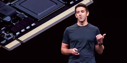 John Ternus speaks on stage, a Mac processor can be seen on the screen behind him