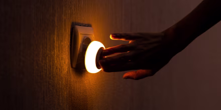 A hand presses a button on a night light plugged into a wall, emitting a warm orange glow