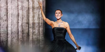Celine Dion raise a hand on stage while performing, wearing a black gown and smiling