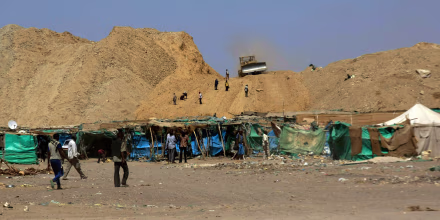 Makeshift shelters lined up against a dry, dusty hillside with a front loader atop it.