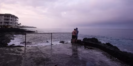 A couple observing a purple sunset over a cloudy sky on the Hawaiian coastline.
