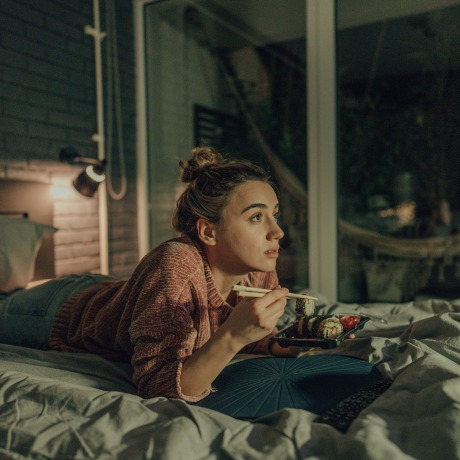 Photo of a young woman watching TV in the bedroom of her apartment; eating sushi and enjoying her night at home alone.