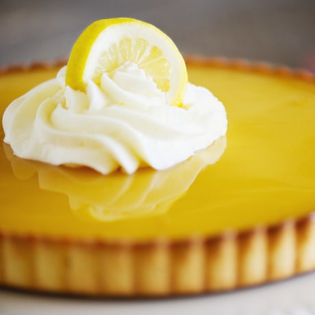 Close-up of lemon tart with whipped cream and lemon slice resting on top of a smooth reflective surface.