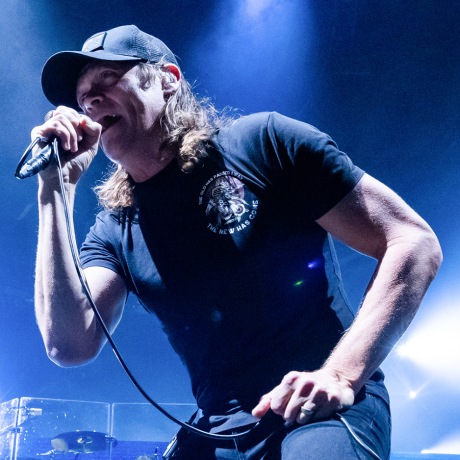 Brad Arnold holds a microphone while singing onstage under blue lights. He's wearing a black t-shirt and black baseball cap.