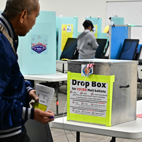 A man holding a voting ballot passes in front of a metal lockbox with a label reading DROP BOX FOR VOTED MAIL BALLOTS inside a polling station.