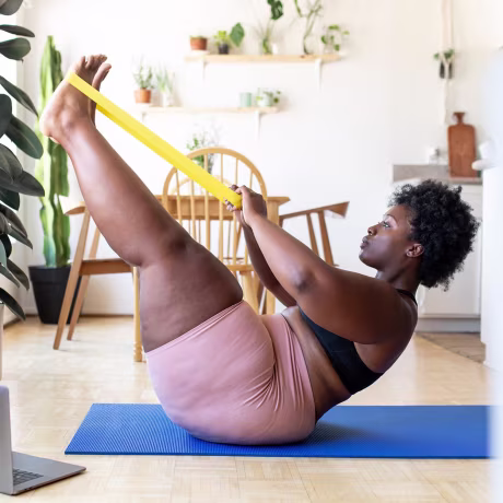Woman sitting on a blue yoga mat, using a yellow resistance band, to workout on her floor