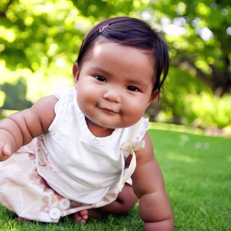 Cute baby girl with a smile on her face, reaching out to take her first crawling step on a lush green lawn at an outdoor park, of mixed ethnic race.