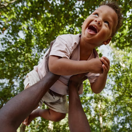 Low angle view at cute baby boy laughing happily while playing with father in park against trees, copy space