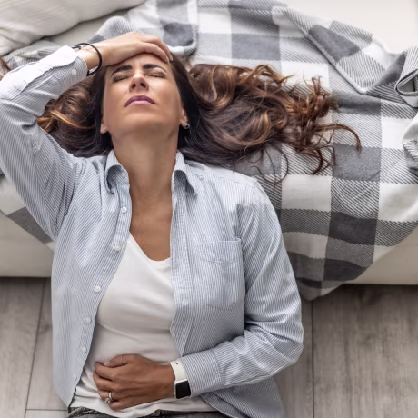 A woman sits on the floor in the living room, leaning her back against a white sofa, and holds her hands on her lower abdomen, suggesting that she is suffering from abdominal pain from menstruation or other health problems.