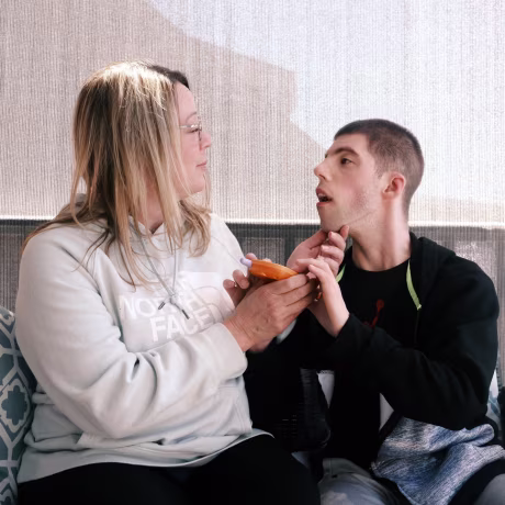 Melissa Gonce sits on a bench on a porch next to Jason, she has her hand on his chin as he holds an orange toy