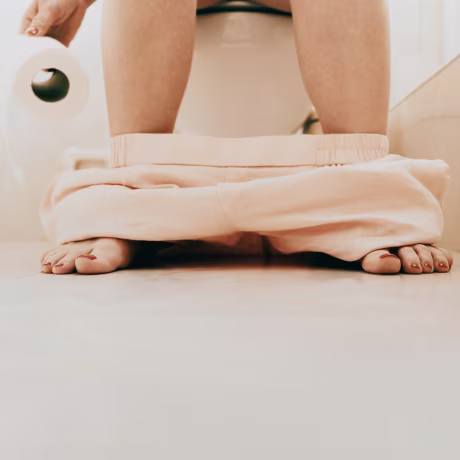 Woman sitting on toilet with pants down holding toilet paper in a bright bathroom