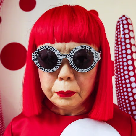 Yayoi Kusama stands in front of her artork, inflated red tubes with white polkadots, she has a bright red bob and sunglasses while wearing a bright read shirt with white polkadots