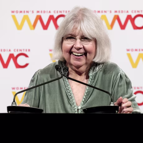 Robin Morgan smiles while standing in front of two microphones on a lectern.