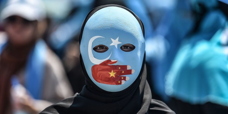 Image: A demonstrator wearing a mask painted with the colours of the flag of East Turkestan and a hand bearing the colours of the Chinese flag