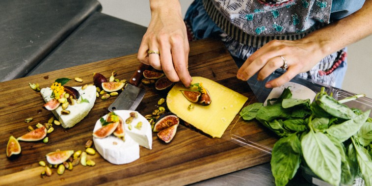 Woman preparing variety cheese plate with figs, nuts, pistachios, basil on wooden chopping board