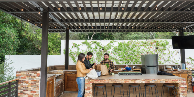 Three people standing in an outdoor backyard kitchen, covered by an overhang, with groceries