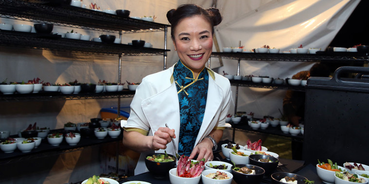 Shirley Chung smiles in a white jacket and blue Chinese-inspired blouse holding a fork over bowls of food