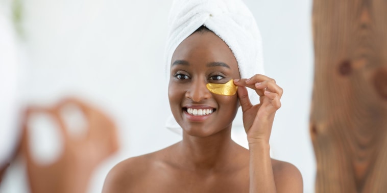 Woman wearing a white towel on her head, putting on gold patches under her eyes, while looking in the mirror
