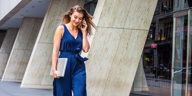 Young Eastern European American Woman talking on cell phone, traveling, working in New York City, wearing blue sleeveless jumpsuit, carrying laptop computer, walking on street outside office building
