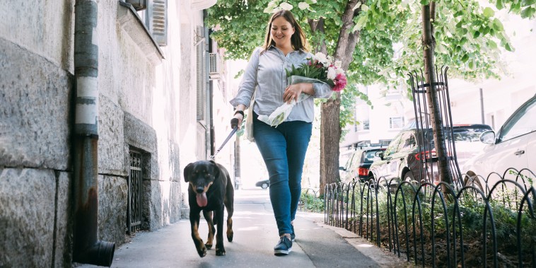 Smiling woman carrying flowers and walking her pet on a sunny day.