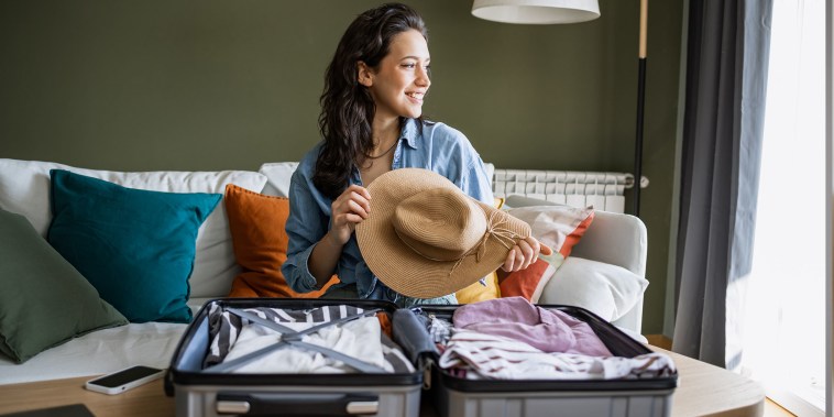 Young woman sitting on the couch and packing suitcase. She is holding a straw hat in her hands